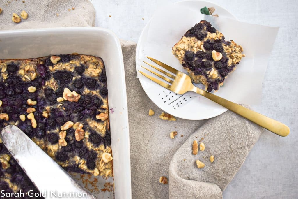 high protein baked oatmeal with blueberries and walnuts in a white dish with one slice cut out and placed on a white round plate with gold fork and tan towel