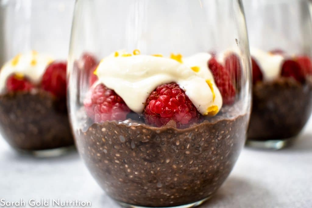 close up of 3 wine glasses filled with dark chocolate chia pudding topped with raspberries and yogurt with meyer lemon zest on grey background. 