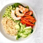 white bowl with shrimp quinoa, edamame, red pepper, cucumber on grey background