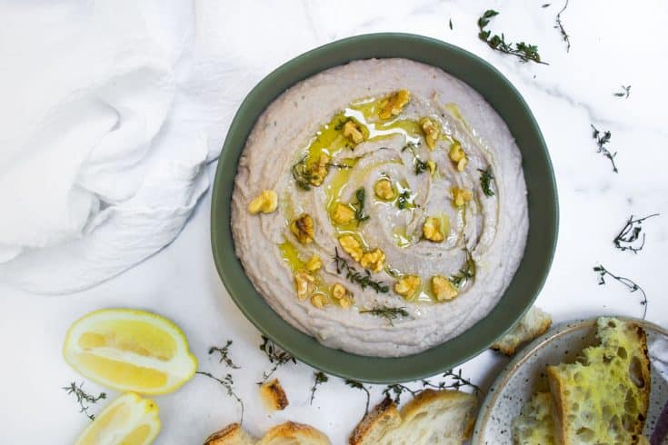 white bean dip in green bowl on marble background with two lemon slices, white towel, and crusty bread scattered.