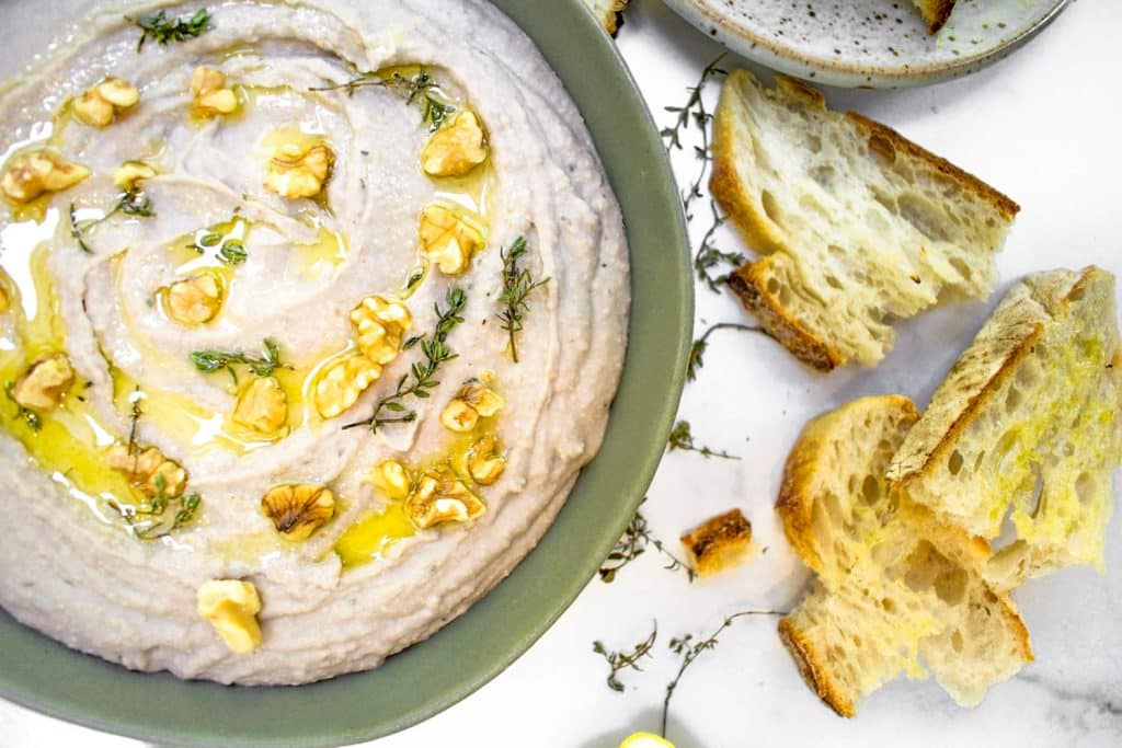 white bean dip in green bowl with walnuts and thyme on top. grey background with crusty bread dipped in olive oil around plate. 