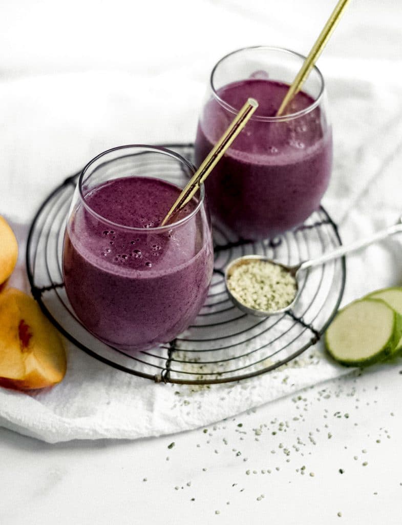 2 blueberry peach smoothies on round trivet with hemp seeds in spoon, and zuchhini and peach slices