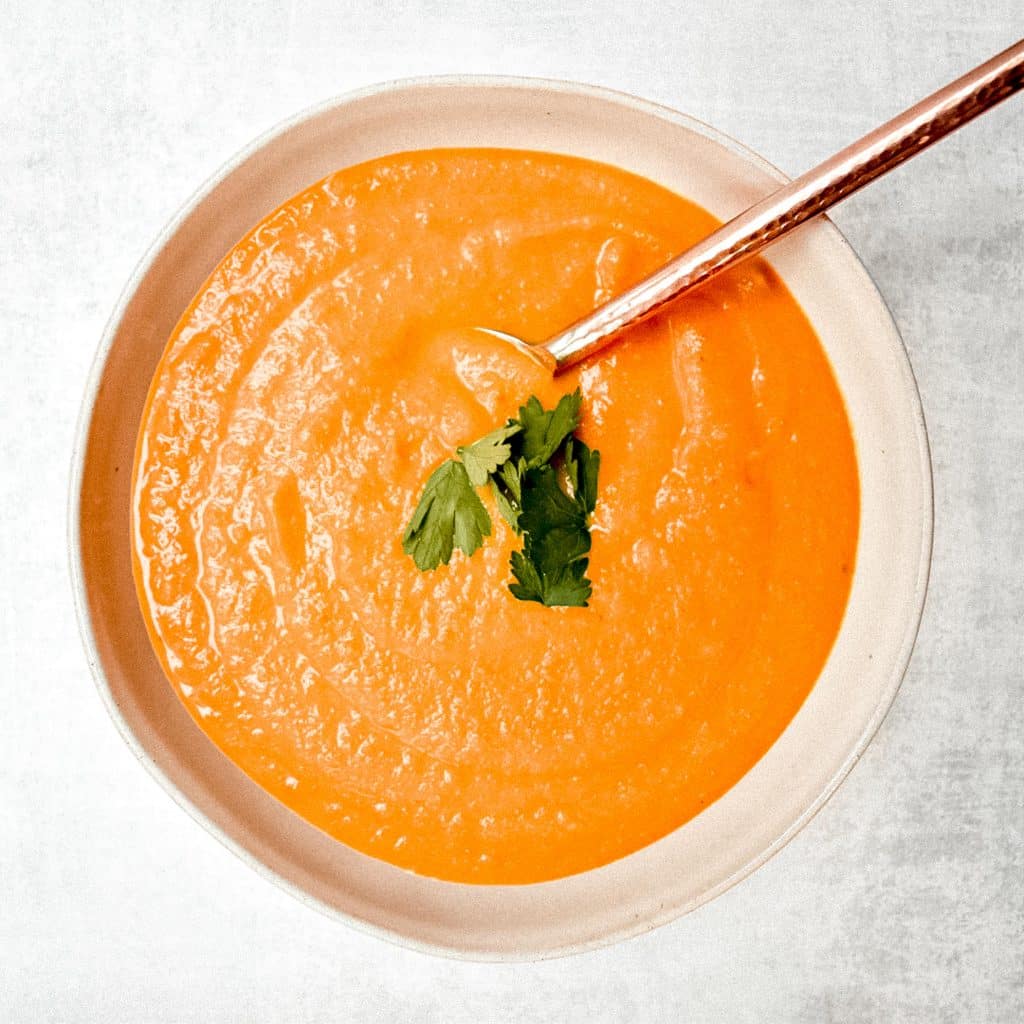 overhead shot of single bowl of carrot ginger soup with carrot leaves on top and a spoon in the bowl