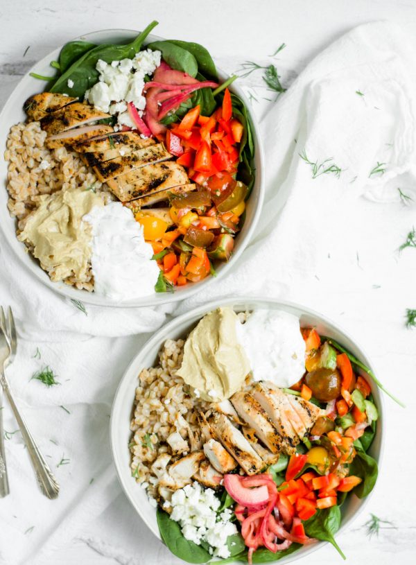 white bowls filled with farro, chicken, veggies, dips, greens, on white background