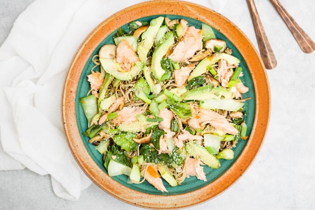 soba noodle salad in large bowl on gray background with white towel and copper serving spoons